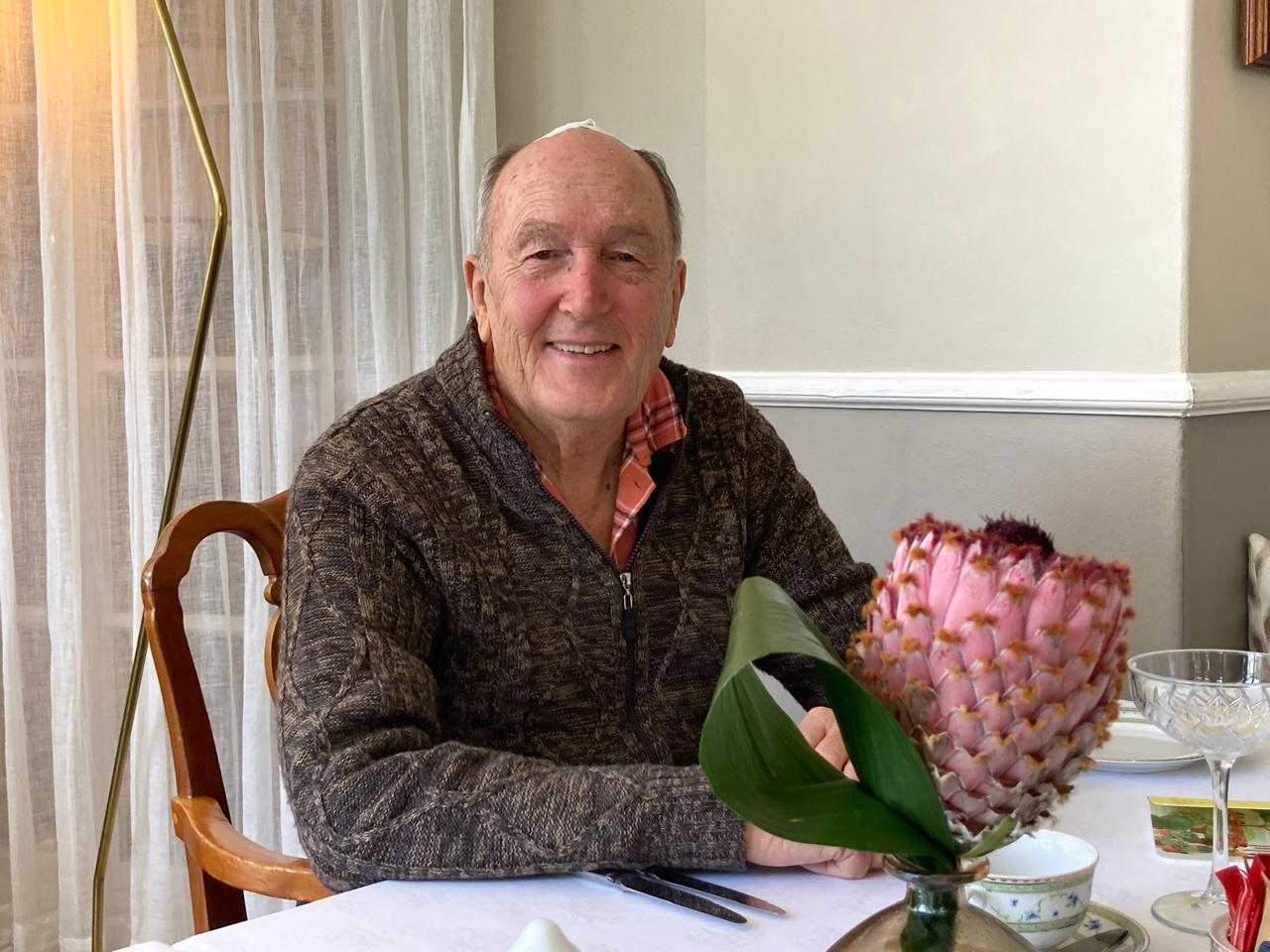 Elderly man smiling, wearing a brown zip-up sweater over a collared shirt, seated at a dining table with a large pink flower in a vase in the foreground.