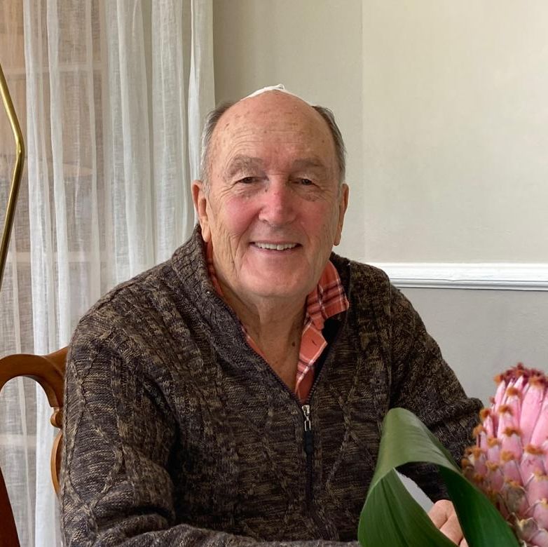 Elderly man smiling, wearing a brown zip-up sweater over a collared shirt, seated at a dining table with a large pink flower in a vase in the foreground.