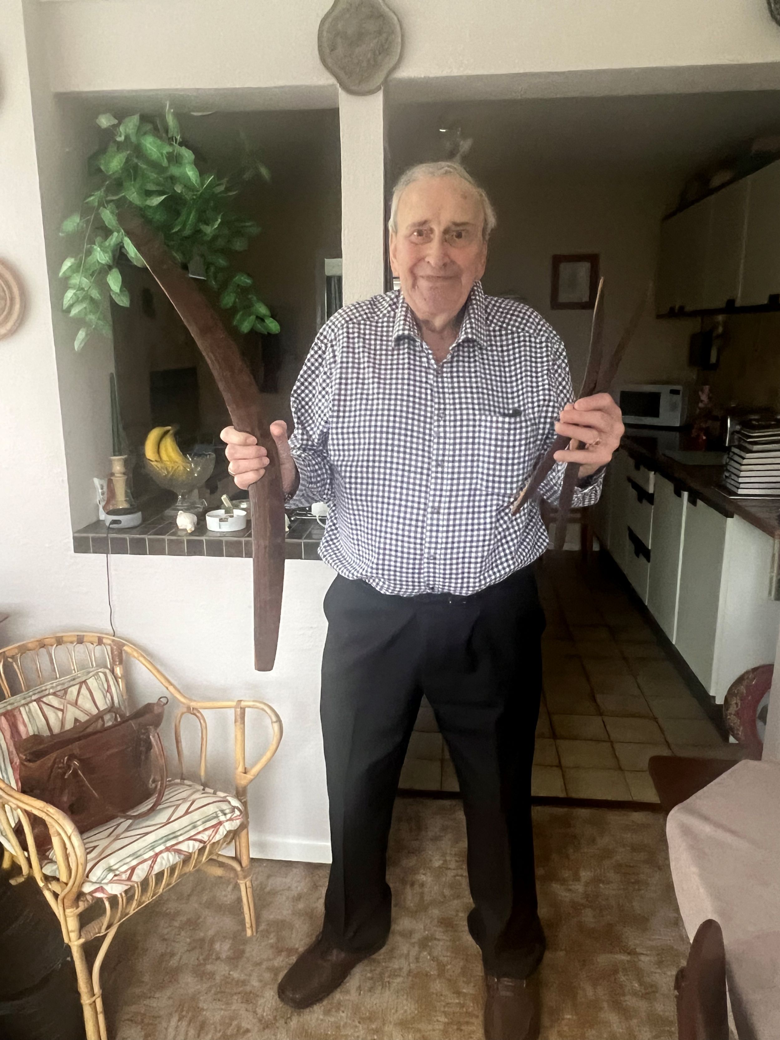 Elderly man smiling indoors, wearing a checked shirt and dark trousers, standing in a kitchen doorway while holding two large wooden boomerangs, one in each hand.