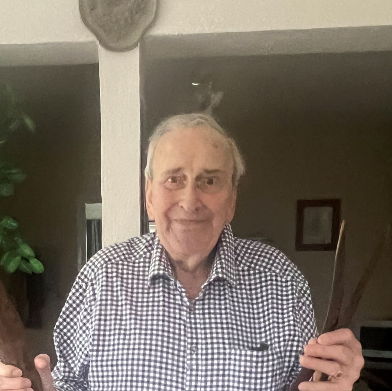 Elderly man smiling indoors, wearing a checked shirt and dark trousers, standing in a kitchen doorway while holding two large wooden boomerangs, one in each hand.