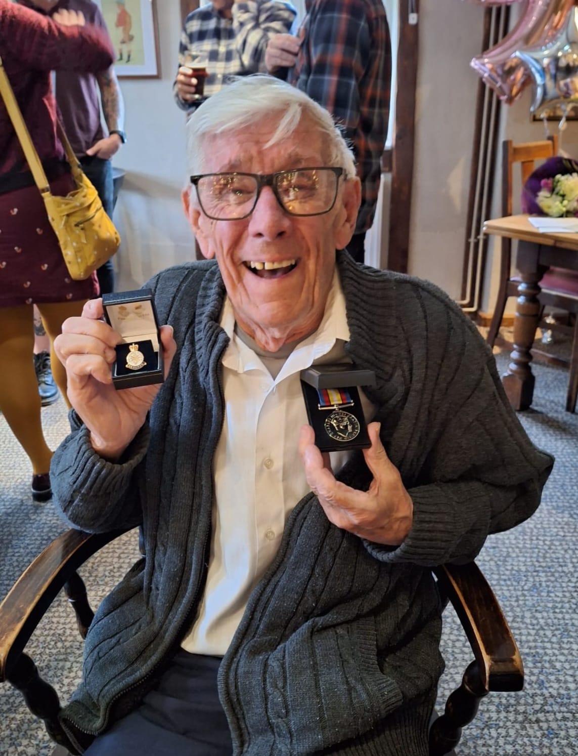 Elderly man smiling broadly while seated, wearing glasses, a white shirt, and a cardigan, proudly holding up two medals in display boxes.