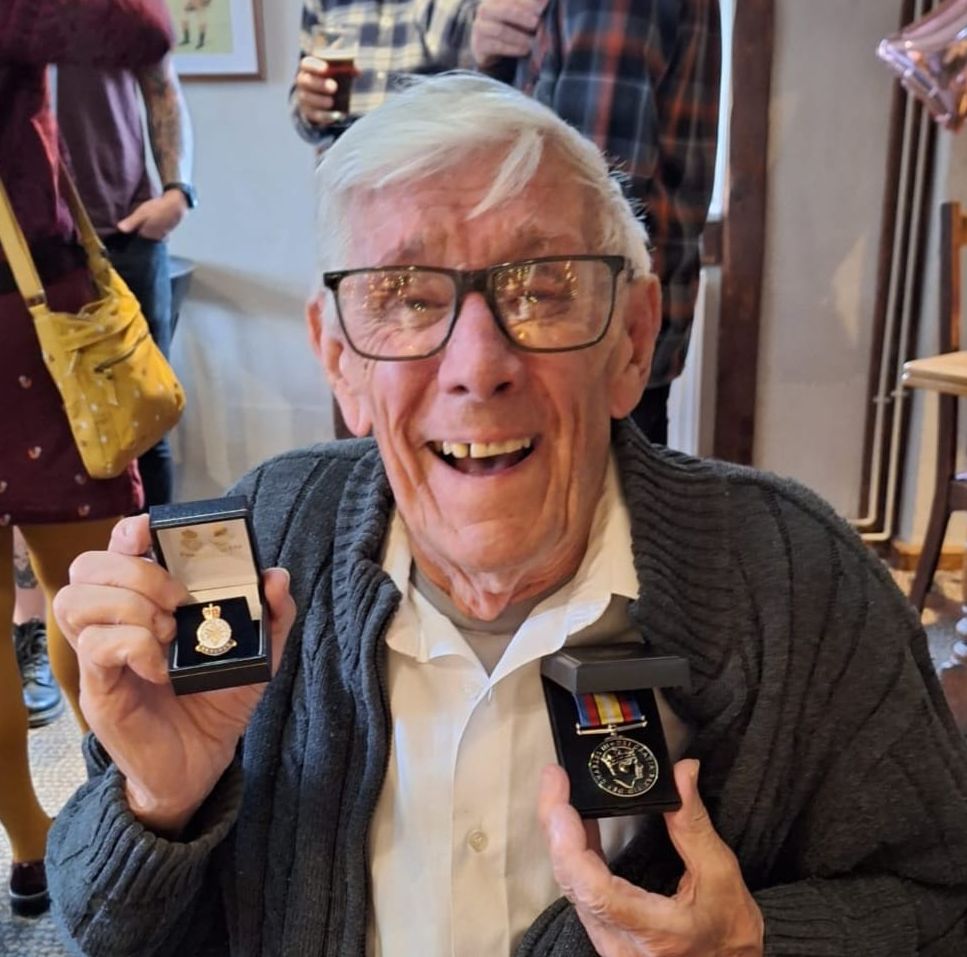 Elderly man smiling broadly while seated, wearing glasses, a white shirt, and a cardigan, proudly holding up two medals in display boxes.
