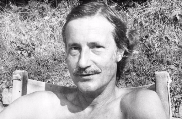 A man with a moustache with his top off, sitting on a garden chair. You can see his head and upper shoulders with grass behind him.