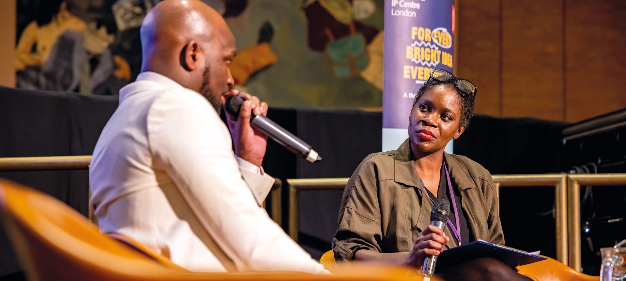 Two business speakers sit on a raised platform with microphones, holding a discussion at an event.