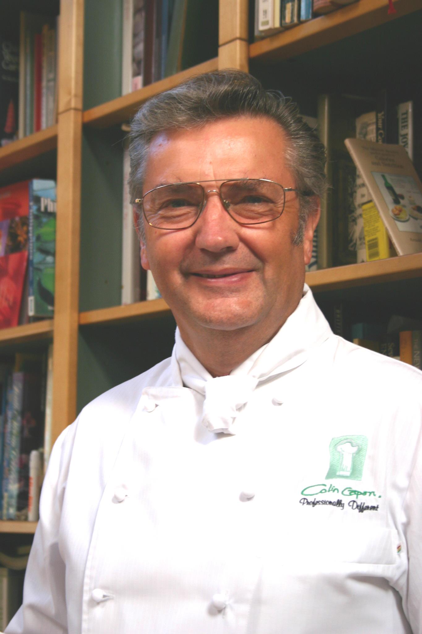 Elderly man smiling, wearing glasses and a white chef’s coat with embroidered text and logo, standing in front of bookshelves.