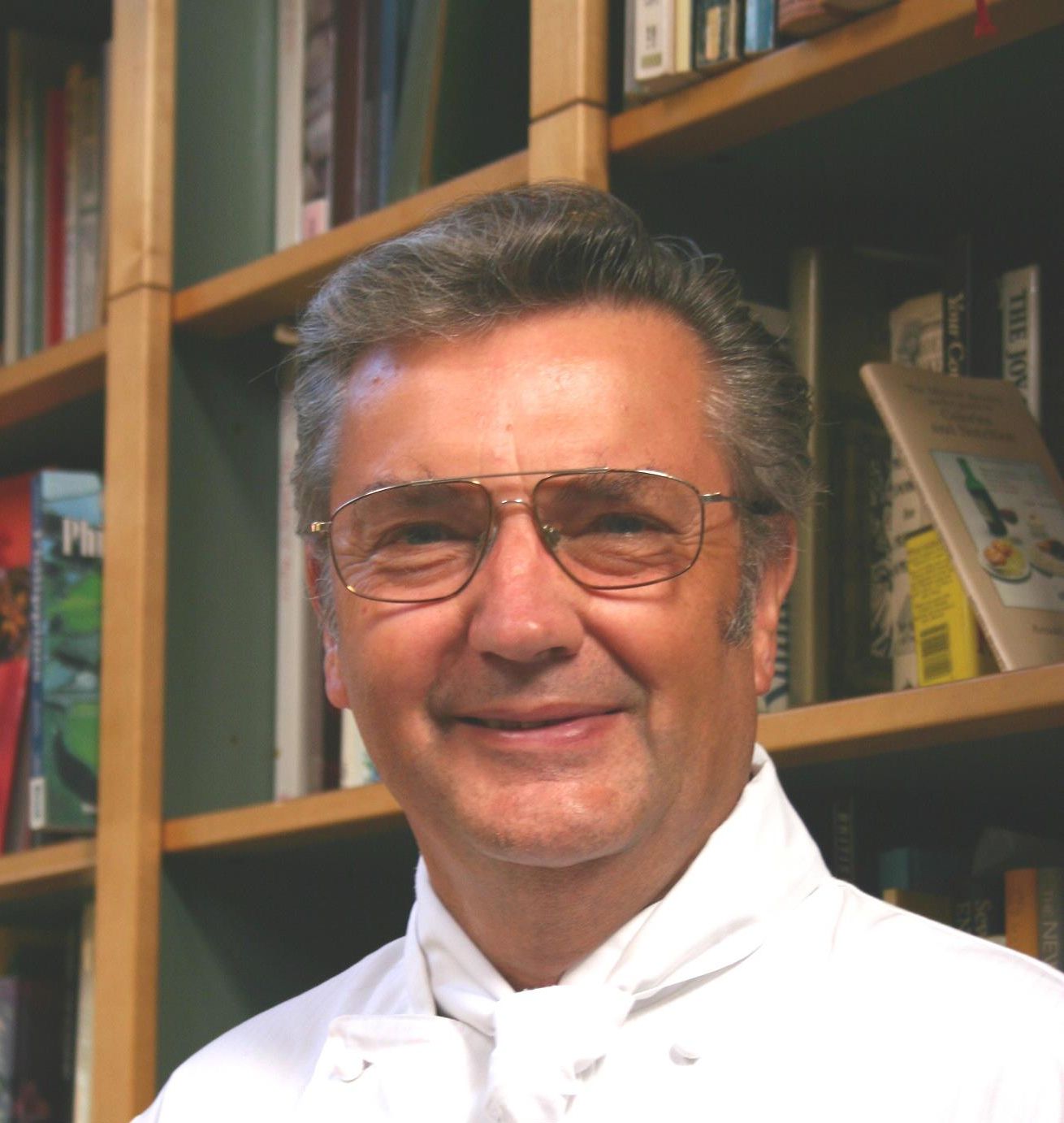 Elderly man smiling, wearing glasses and a white chef’s coat with embroidered text and logo, standing in front of bookshelves.