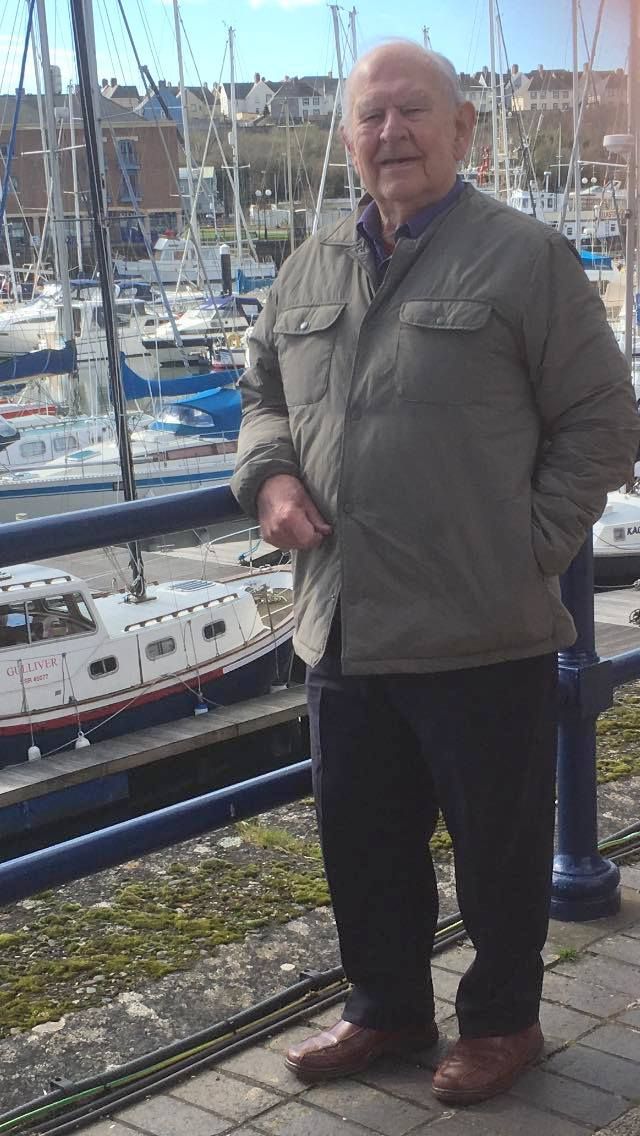 Elderly man standing by a marina, wearing a gray jacket, dark trousers, and brown shoes, with moored sailboats and buildings in the background.