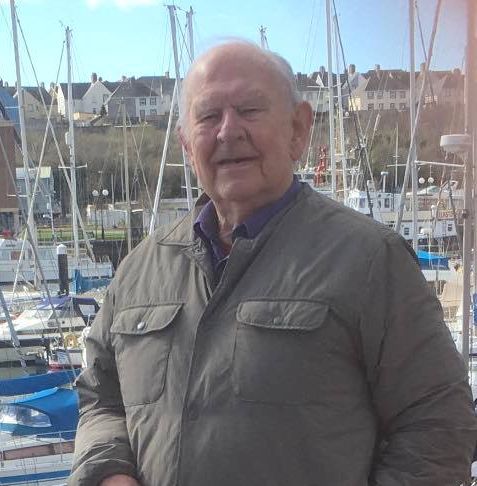 Elderly man standing by a marina, wearing a gray jacket, dark trousers, and brown shoes, with moored sailboats and buildings in the background.
