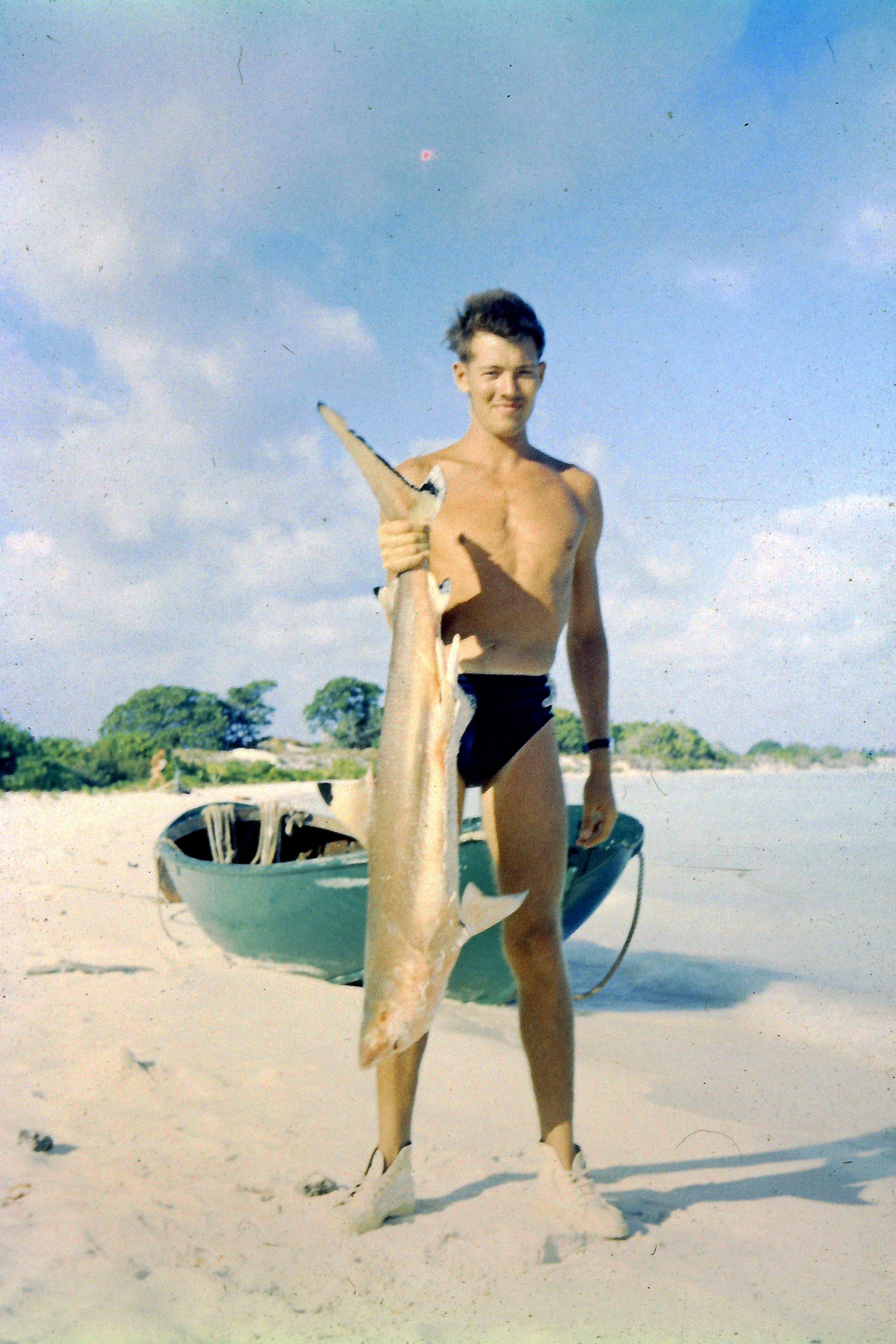 Sapper Frank Bools holds up a fish just over half the size of his six foot frame. He stands on a white beach with a small fishing boat in the background.