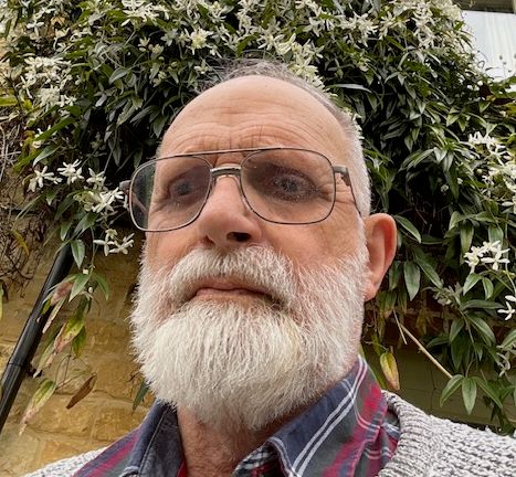 Elderly man with a white beard and glasses, wearing a grey knit sweater over a red and blue checked shirt, looking serious in front of a wall covered with flowering vines.