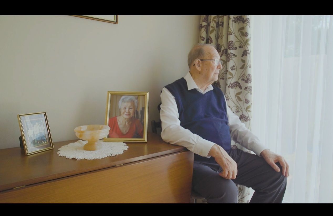 An elderly man sits by a window, gazing outside with a thoughtful expression. He wears glasses, a white shirt, and a navy sweater vest. Beside him on a wooden cabinet are framed photographs, including a portrait of an elderly woman in a red top, and a smaller family photo. A decorative bowl on a lace doily is also on the cabinet. Floral curtains frame the window, letting in soft daylight