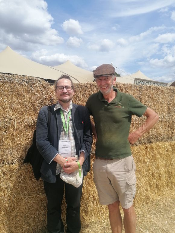Two men standing in front of a wall of hay bales. One is older in shorts and t shirt with a flat cap, the other is younger in a casual jacket and shirt, with a lanyard around his neck.