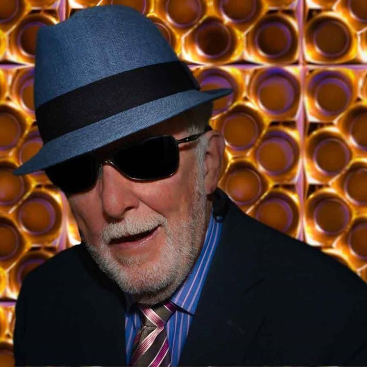 Elderly man with a white beard smiling, wearing a blue fedora, dark sunglasses, striped shirt, and pink patterned tie under a dark blazer, standing in front of a brightly lit patterned background.