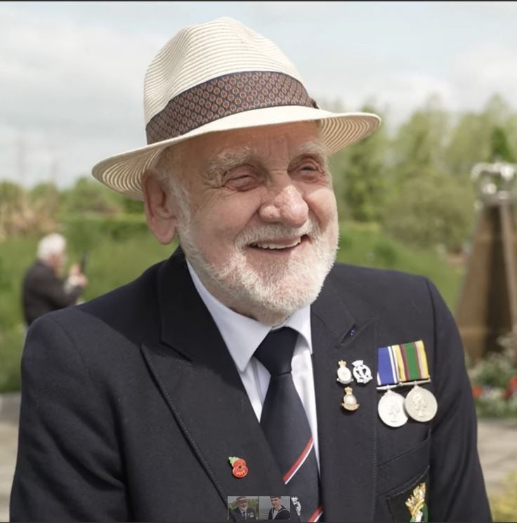 Elderly man with a white beard smiling outdoors, wearing a light-colored hat, dark blazer, and tie, decorated with several military medals and pins.