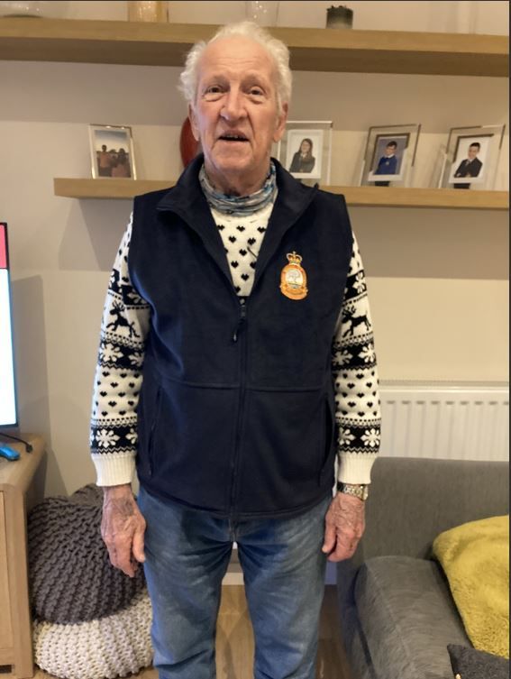 Elderly man standing indoors, wearing a patterned sweater with reindeer motifs under a dark vest featuring an embroidered crest badge, with framed family photos on a shelf behind him.