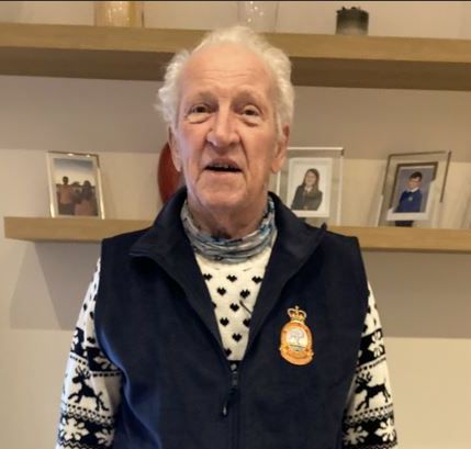 Elderly man standing indoors, wearing a patterned sweater with reindeer motifs under a dark vest featuring an embroidered crest badge, with framed family photos on a shelf behind him.
