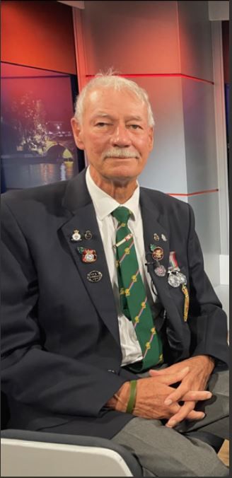 Elderly man with white hair and mustache, wearing a dark blazer decorated with multiple medals and pins, a white shirt, and a green patterned tie, seated in a studio with hands folded and a calm expression.