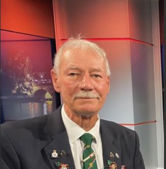 Elderly man with white hair and mustache, wearing a dark blazer decorated with multiple medals and pins, a white shirt, and a green patterned tie, seated in a studio with hands folded and a calm expression.