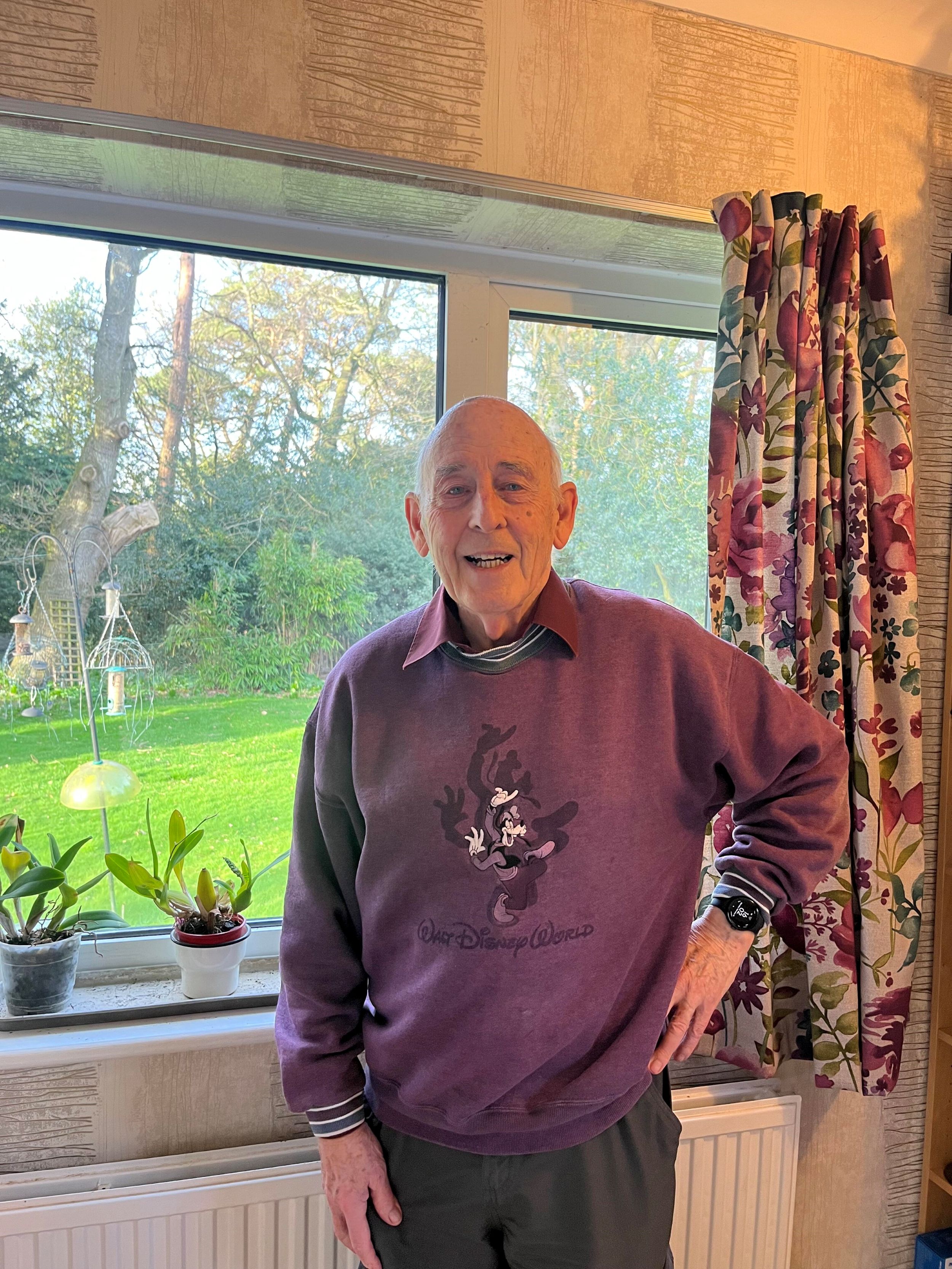 Elderly man smiling indoors, wearing a purple Walt Disney World sweatshirt, standing in front of a large window with a garden view and floral curtains.