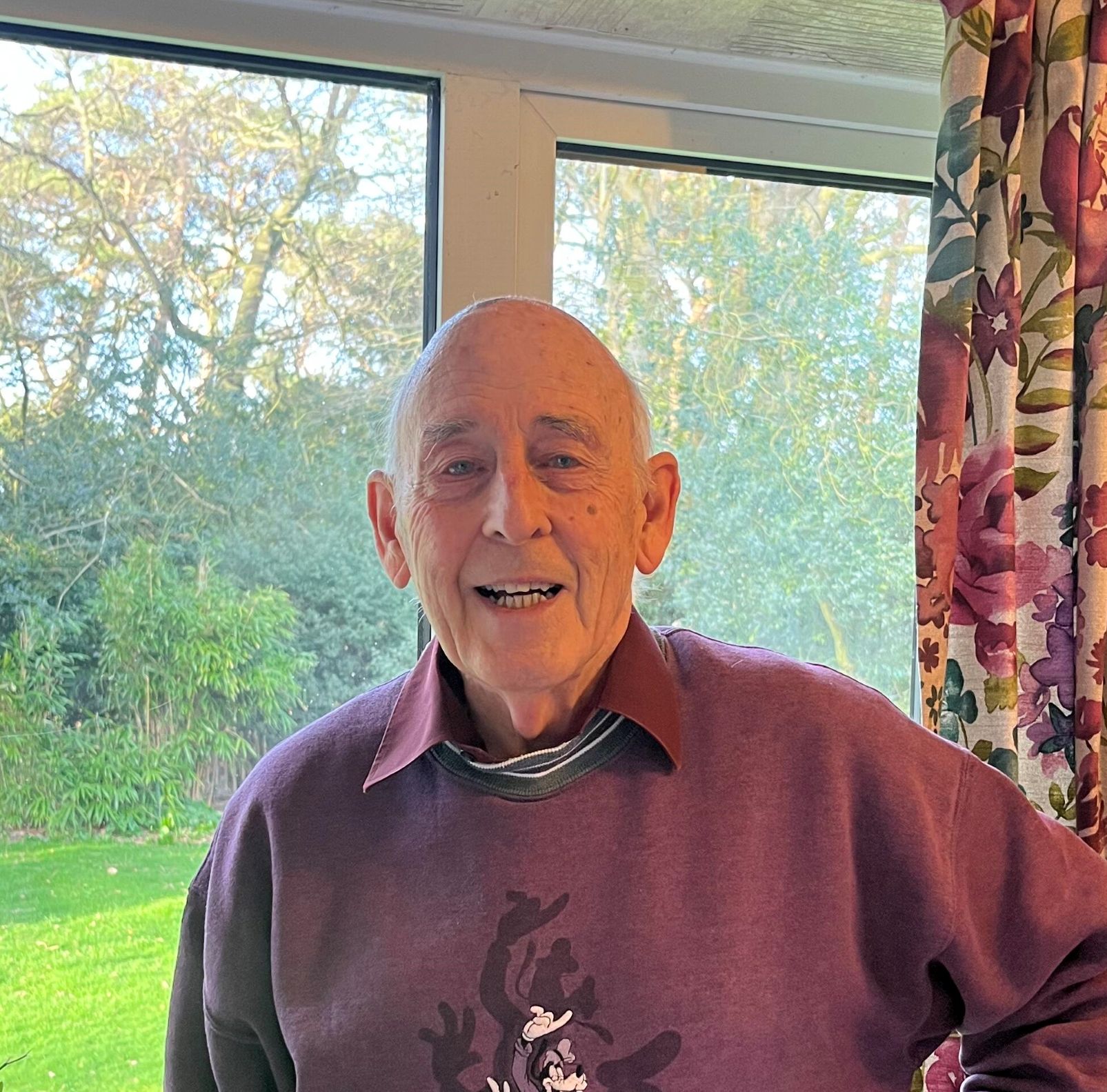 Elderly man smiling indoors, wearing a purple Walt Disney World sweatshirt, standing in front of a large window with a garden view and floral curtains.