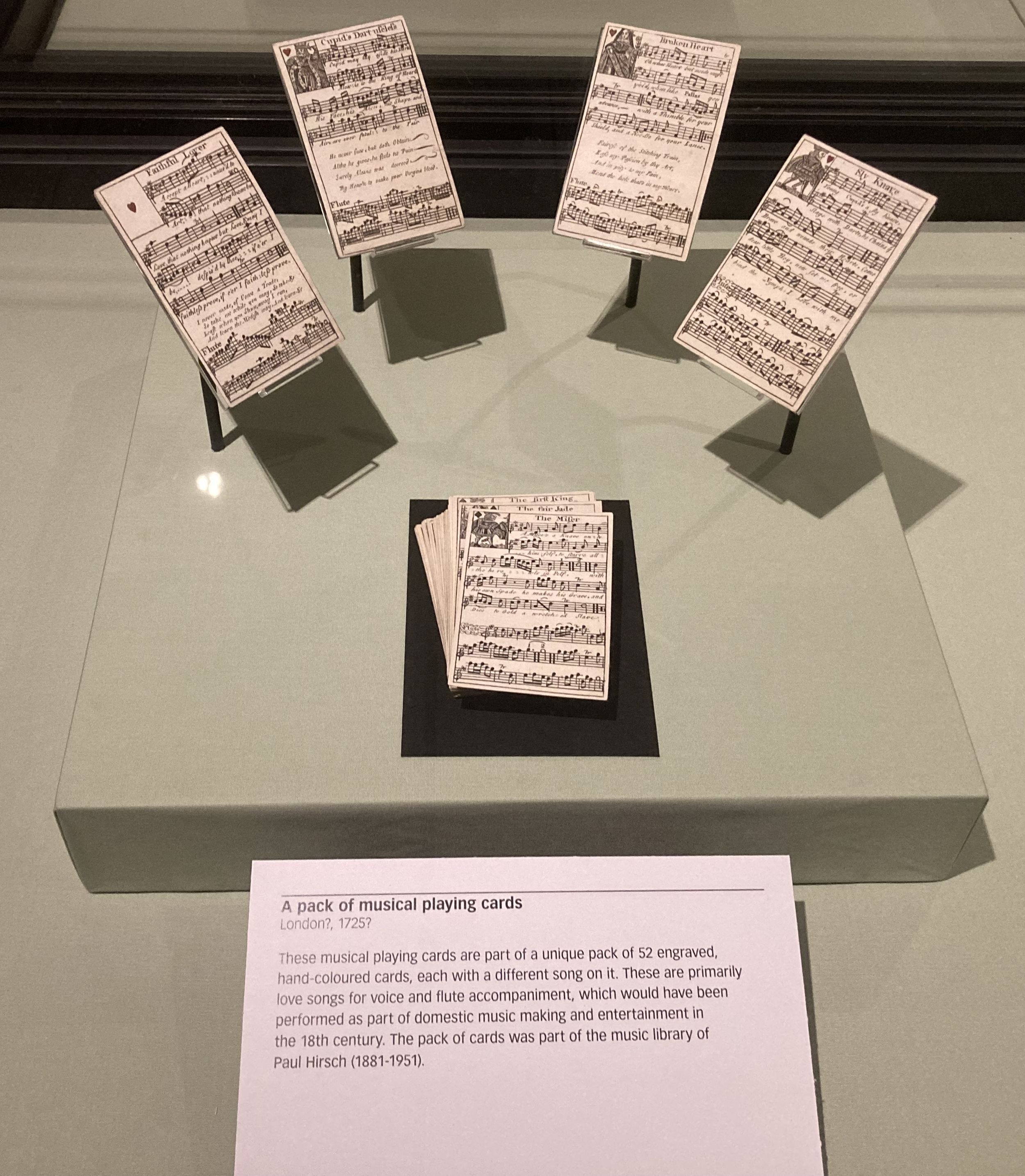 Musical playing cards on display in an exhibition cabinet. The description panel says: "These musical playing cards are a unique pack of 52 engraved, hand-coloured cards, each with a different song on it. These are primarily love songs for voice and flute accompaniment, which would have been performed as part of domestic music making and entertainment in the 18th centure. The pack of cards was part of the music library of Paul Hirsch (1881-1951)."
