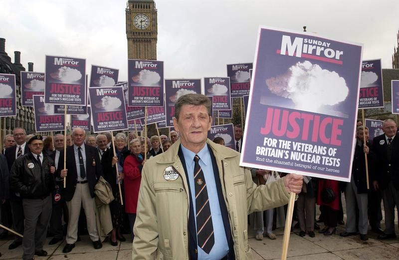 Ken McGinley in a suit and tie, holding up a placard sponsored by the Sunday Mirror and featuring the words: 'Justice for the Veterans of Britain's Nuclear Tests'. Big Ben is in the background and McGinley is surrounded by other veterans in suits.