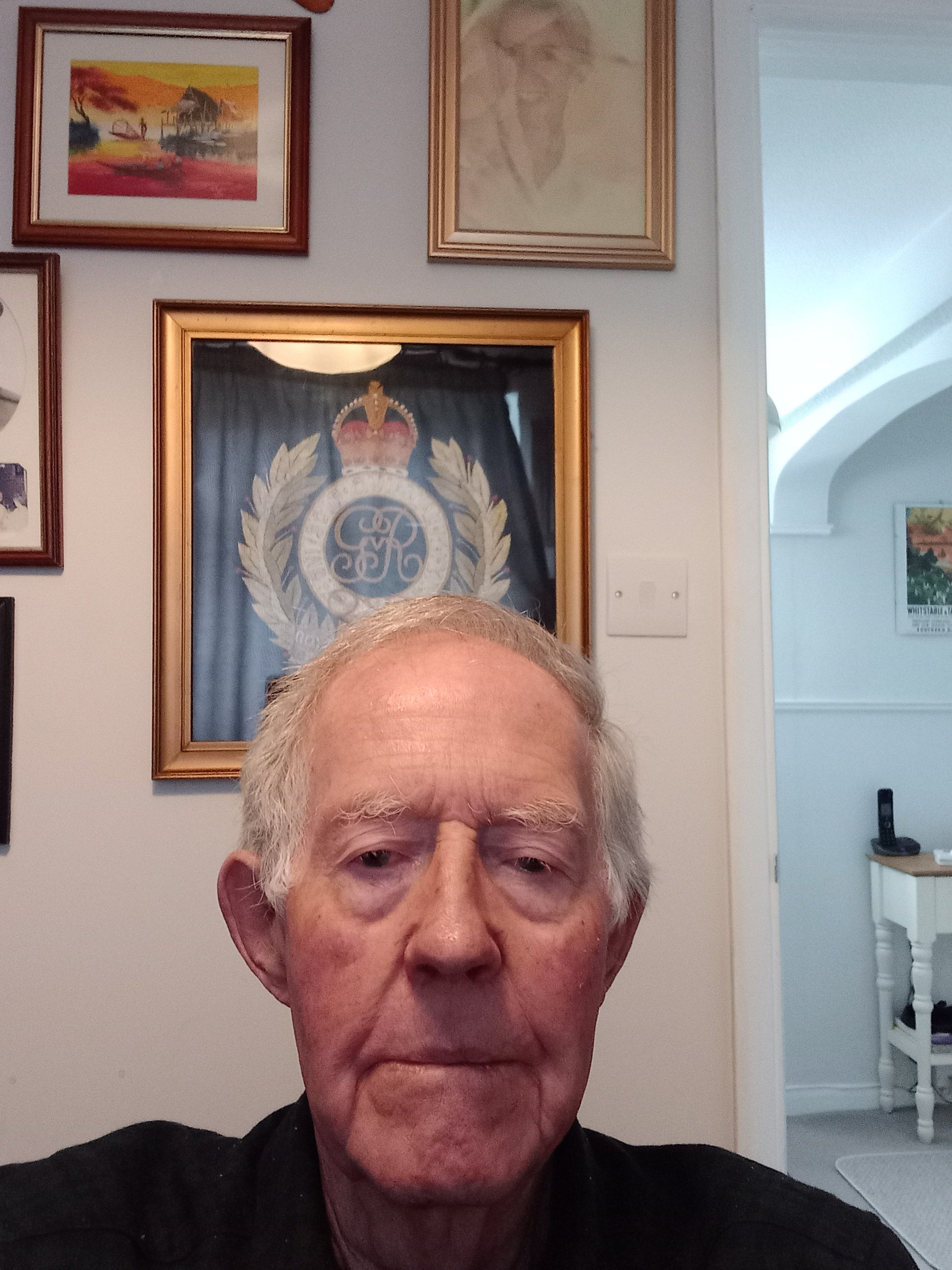 Elderly man with short gray hair and a serious expression, seated in a room with framed artwork and a large crest with a crown displayed on the wall behind him.