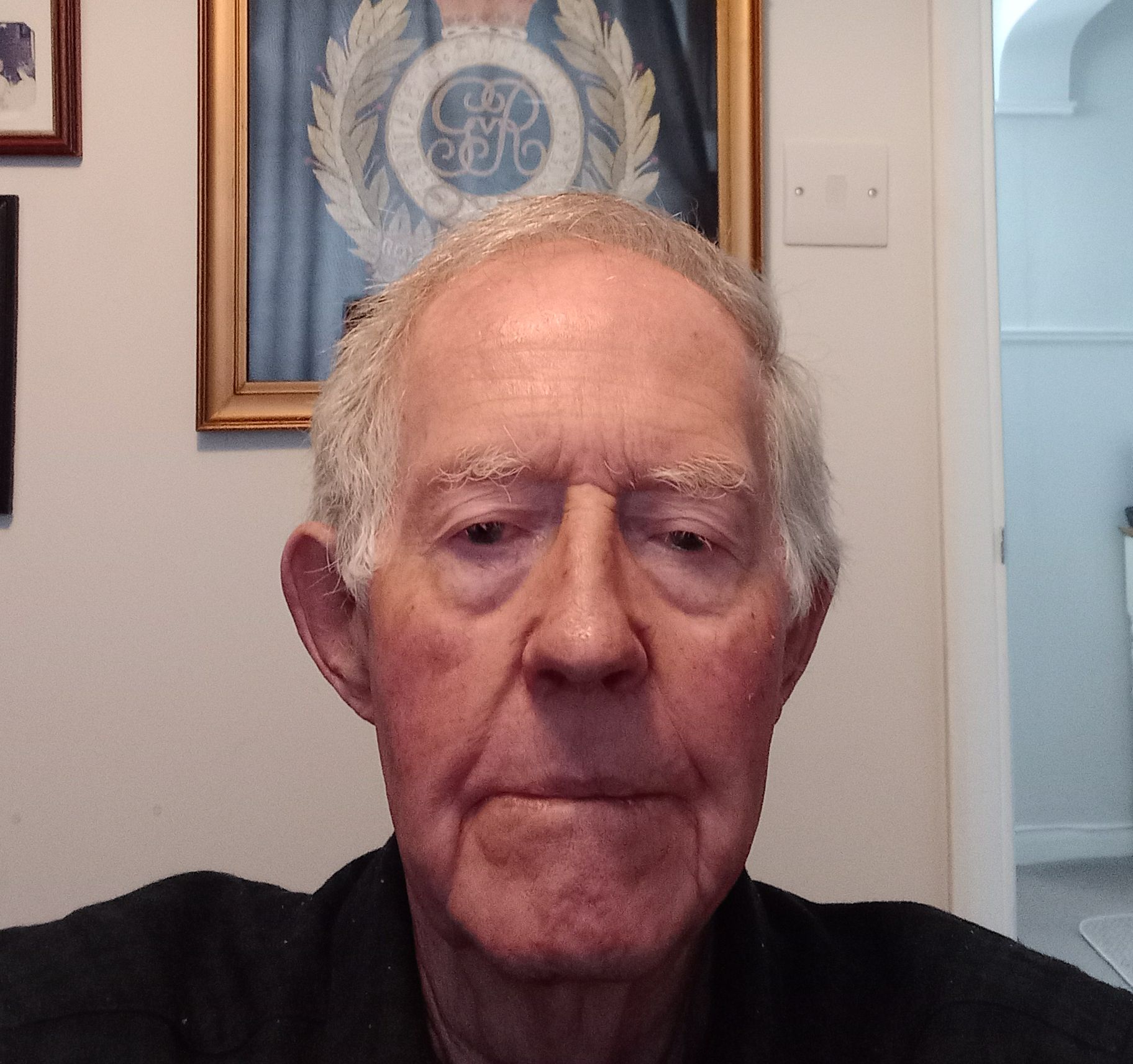 Elderly man with short gray hair and a serious expression, seated in a room with framed artwork and a large crest with a crown displayed on the wall behind him.