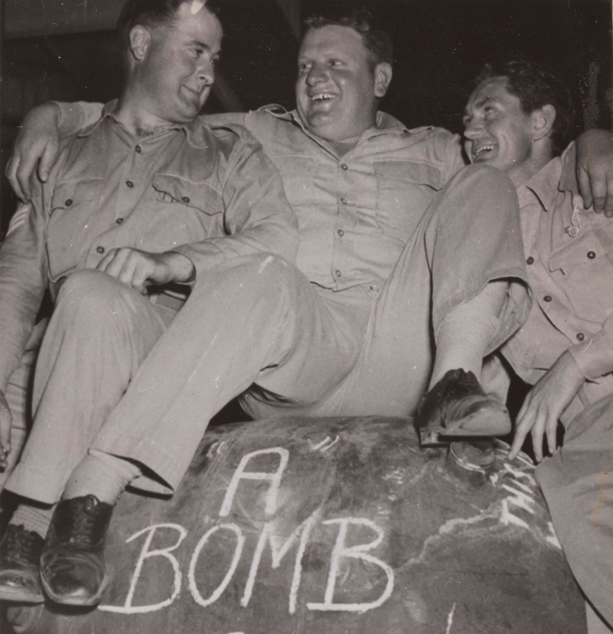 Three soldiers in khaki sit on top of a large deloused mine, with the words 'A-Bomb Stand Clear' daubed on it.