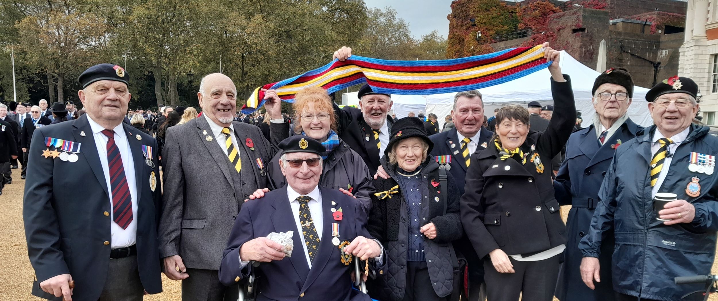Seven veterans and three of their wives pose for a photograph to celebrate wearing the Nuclear Test Medal. They are holding up a multi-coloured scarf, which is designed to replicate the colours of the medal ribbon, which themselves were selected to symbolise a British nuclear bomb test.