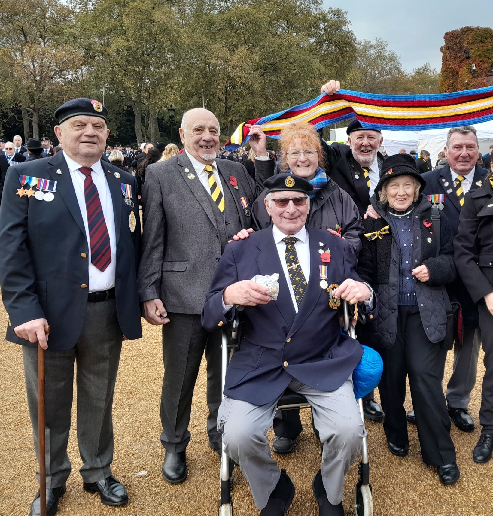 Seven veterans and three of their wives pose for a photograph to celebrate wearing the Nuclear Test Medal. They are holding up a multi-coloured scarf, which is designed to replicate the colours of the medal ribbon, which themselves were selected to symbolise a British nuclear bomb test.