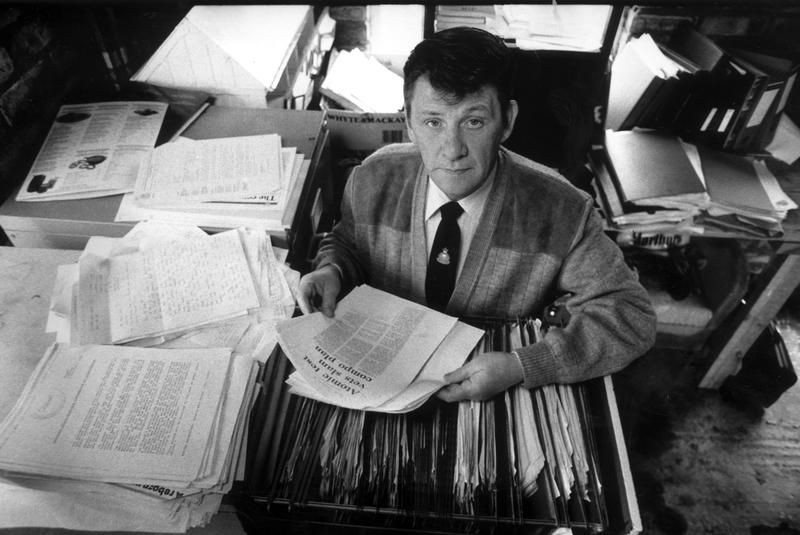 Ken McGinley looks up at the camera with a document in his hand whilst sat at a desk.