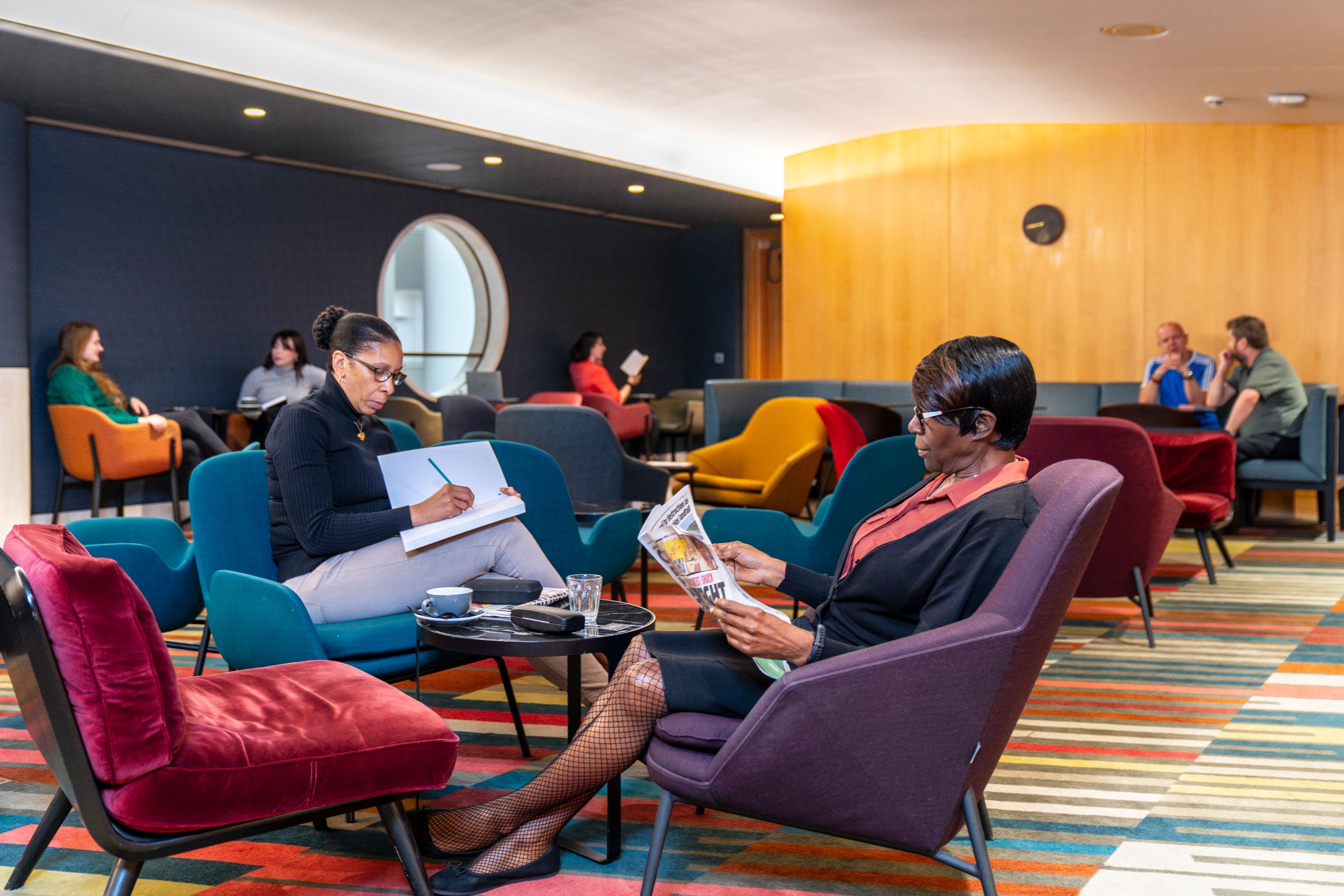 Several people sit talking and reading papers and books in the Members' Room of the British Library, London.