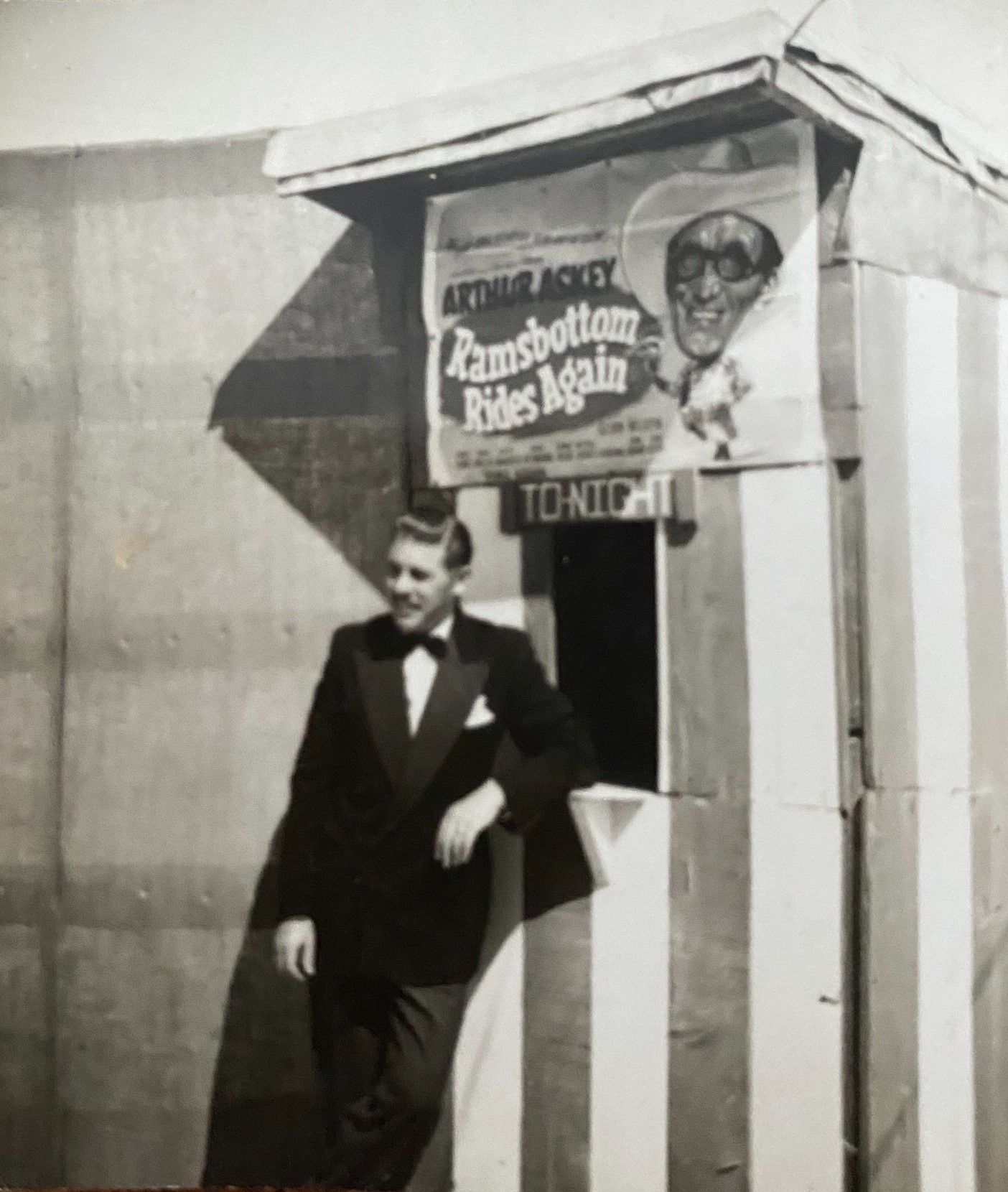 Black-and-white photo of a young man in a tuxedo with a bow tie, leaning casually against a striped booth. Above him hangs a poster advertising “Arthur Askey – Ramsbottom Rides Again” with the word “Tonight” displayed below it.