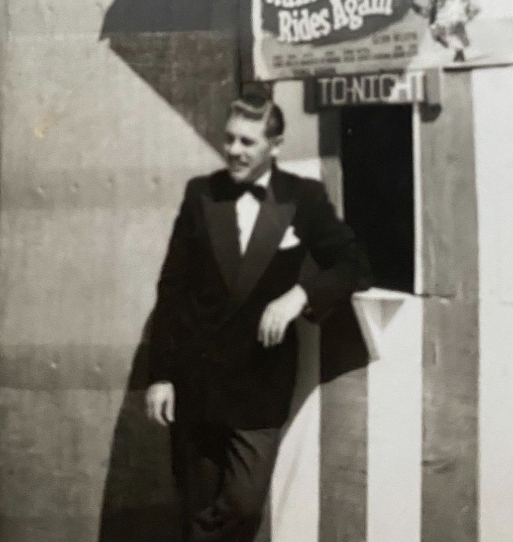 Black-and-white photo of a young man in a tuxedo with a bow tie, leaning casually against a striped booth. Above him hangs a poster advertising “Arthur Askey – Ramsbottom Rides Again” with the word “Tonight” displayed below it.