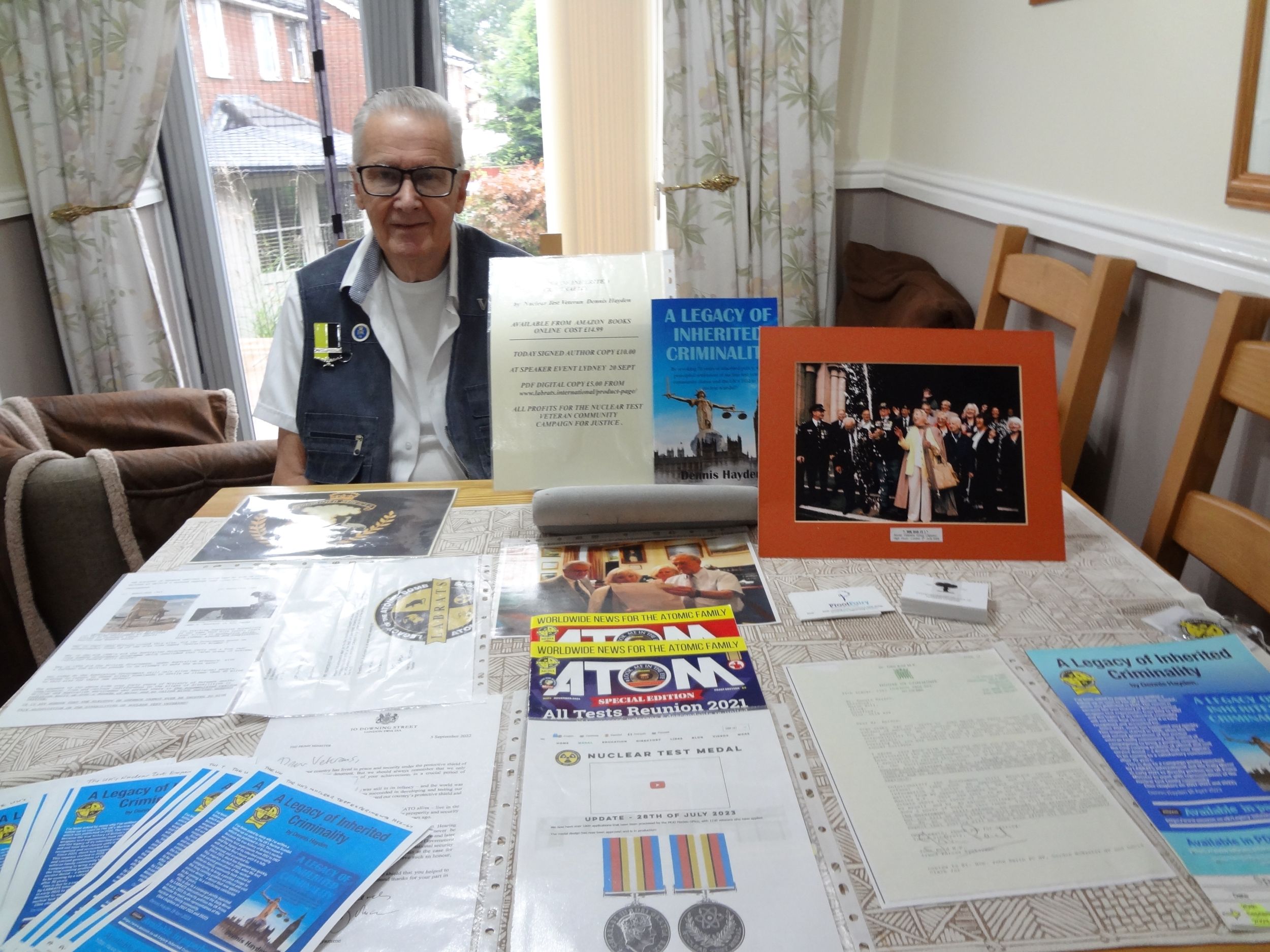 Elderly man wearing glasses and a vest with a medal pin, seated at a table covered with memorabilia including documents, booklets, photographs, and a framed picture of a formal ceremony.