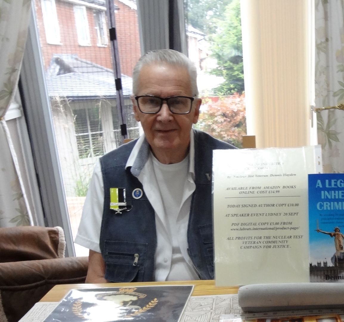 Elderly man wearing glasses and a vest with a medal pin, seated at a table covered with memorabilia including documents, booklets, photographs, and a framed picture of a formal ceremony.
