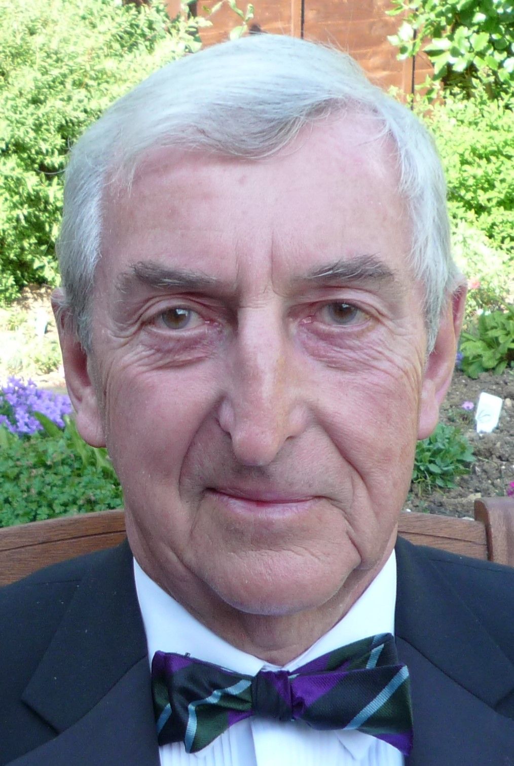 Elderly man with gray hair wearing a tuxedo with a colorful bow tie, looking directly at the camera with a composed expression, with greenery and flowers in the background.
