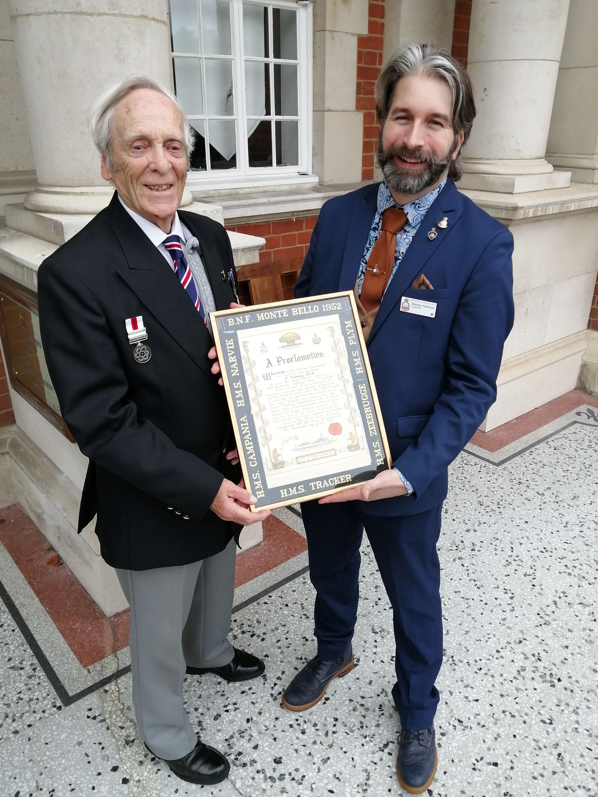 Elderly man in a blazer with medals, smiling as he stands beside a younger man in a suit. They are both holding a framed proclamation, posed outside a building with columns and red brick walls.