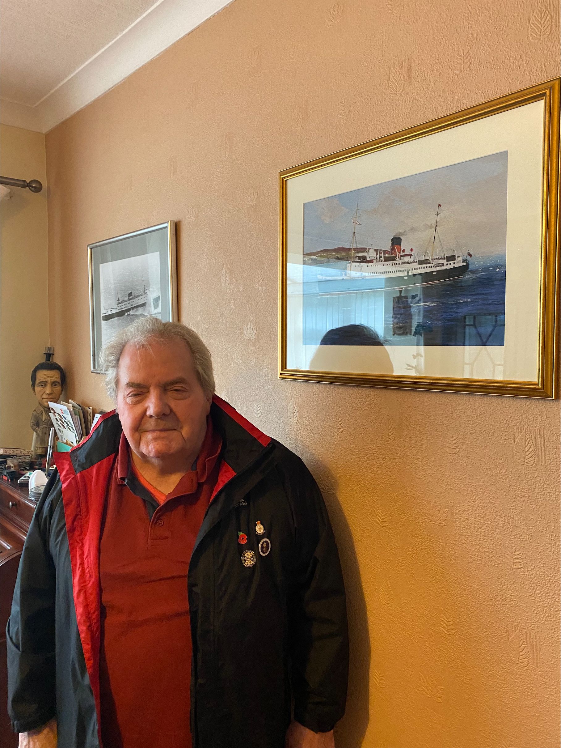 Elderly man wearing a red shirt and black jacket with pins, standing indoors beside framed paintings of ships on a wall.