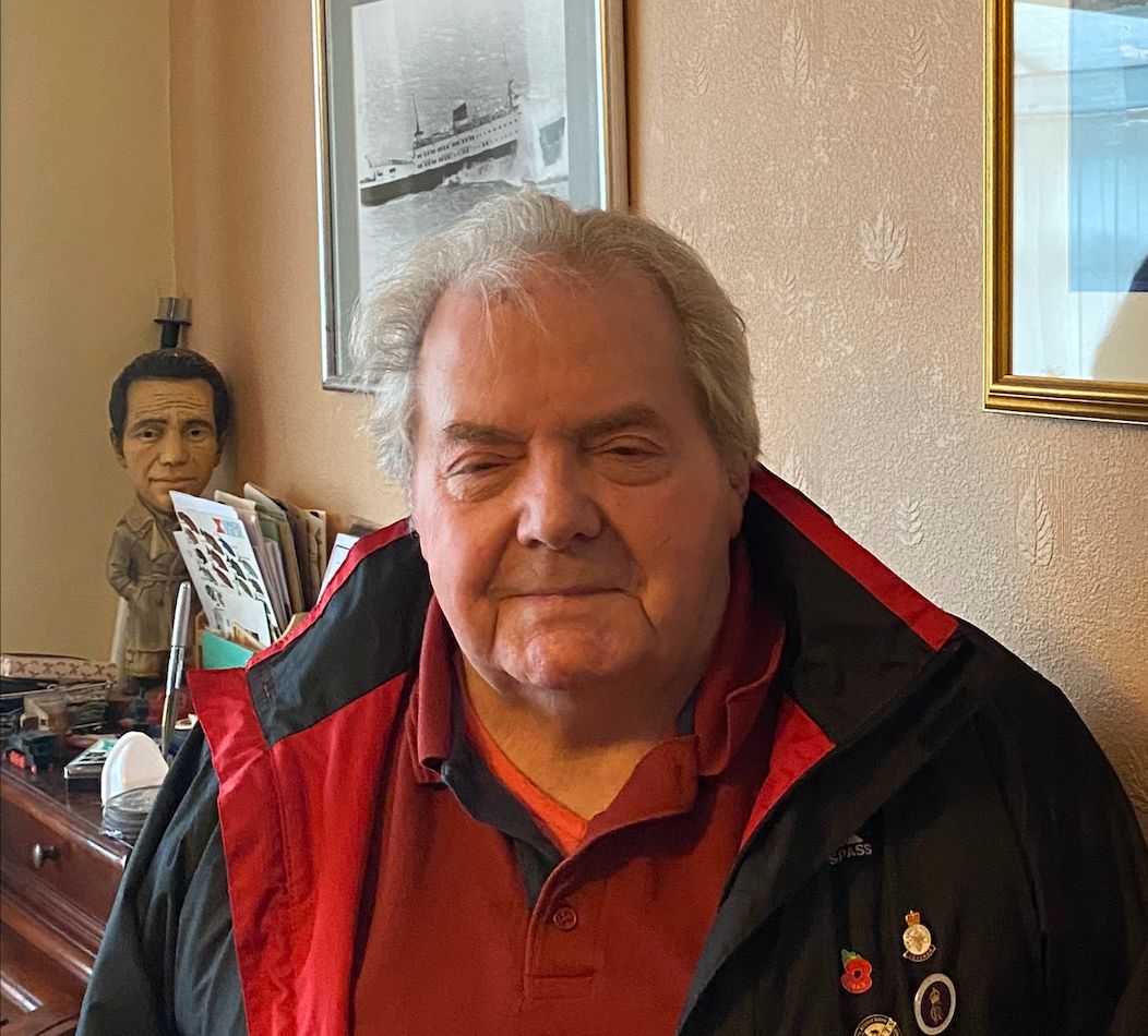 Elderly man wearing a red shirt and black jacket with pins, standing indoors beside framed paintings of ships on a wall.