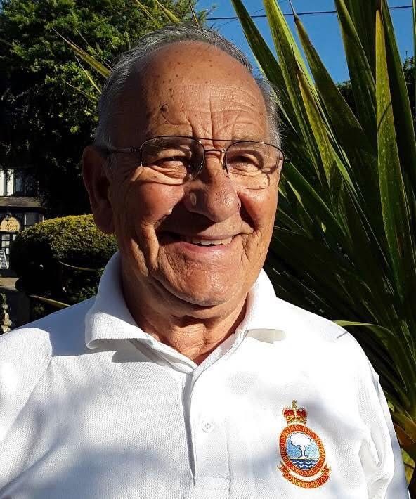 Elderly man smiling, wearing glasses and a white polo shirt with an embroidered crest badge, standing in front of greenery.