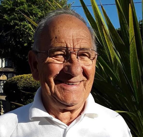 Elderly man smiling, wearing glasses and a white polo shirt with an embroidered crest badge, standing in front of greenery.