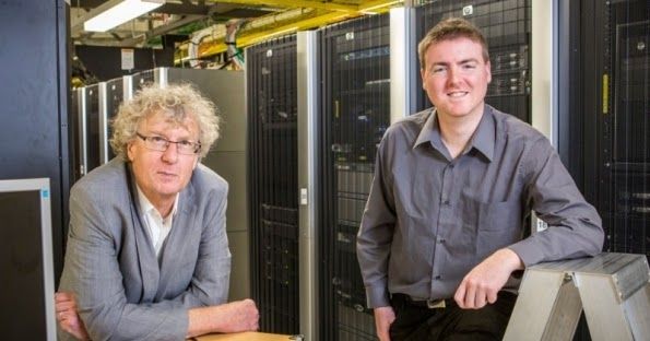 Two men in a server room. One with curly gray hair and glasses is leaning on a desk, while the other, younger man in a gray shirt, is smiling and leaning on a metal step ladder. Behind them are tall racks of computer servers and cables.