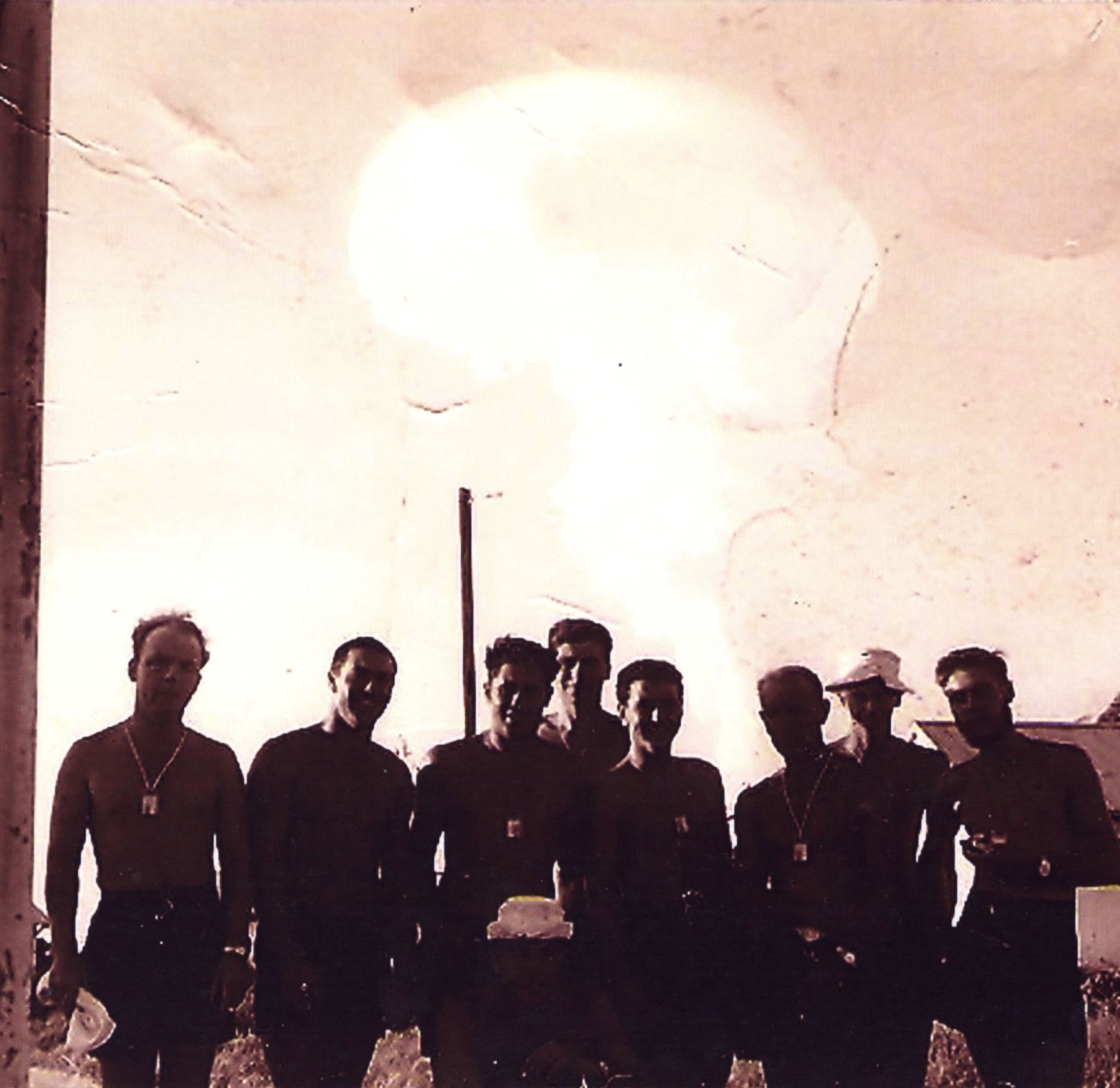 Eight men stand in front of the bright light of a mushroom cloud.