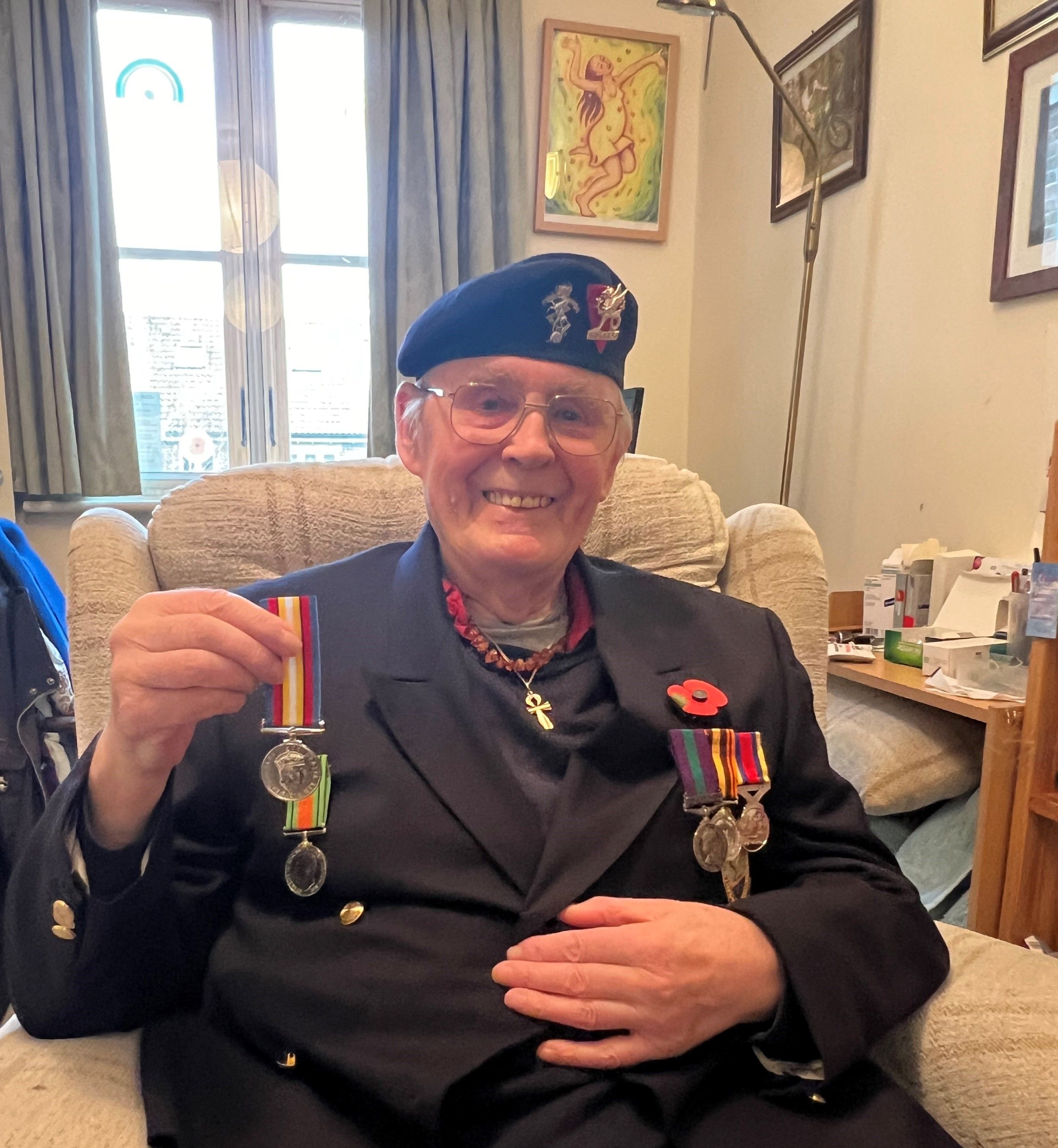 Elderly man smiling while seated, wearing glasses, a dark blazer with medals and a red poppy pin, and a beret with insignia, proudly holding up a medal in his hand.