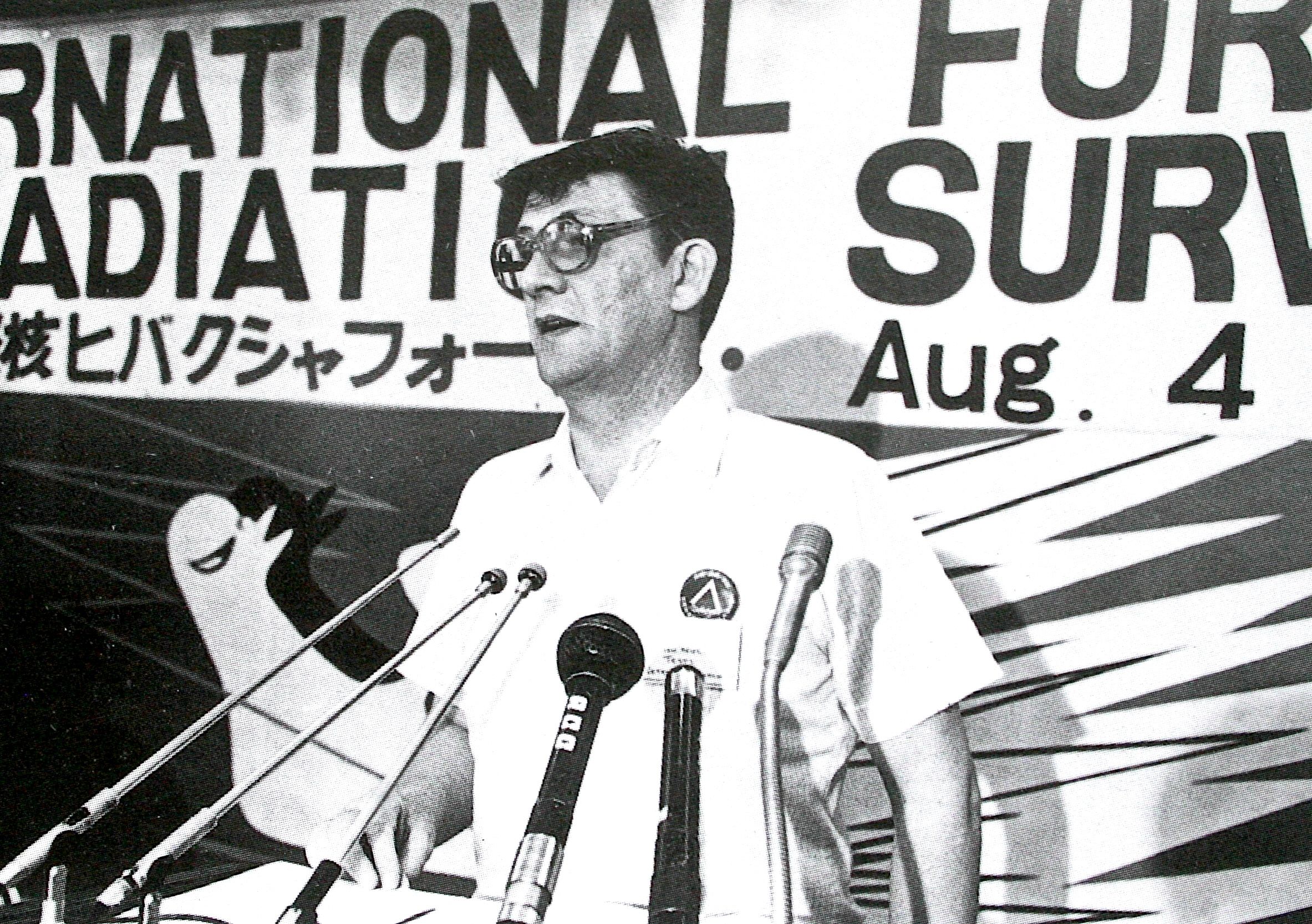 Ken McGinley sat a table wearing glasses with microphones in front of him. A banner behind him contains Japanese script and the date '4 August', which is Hiroshima Day.