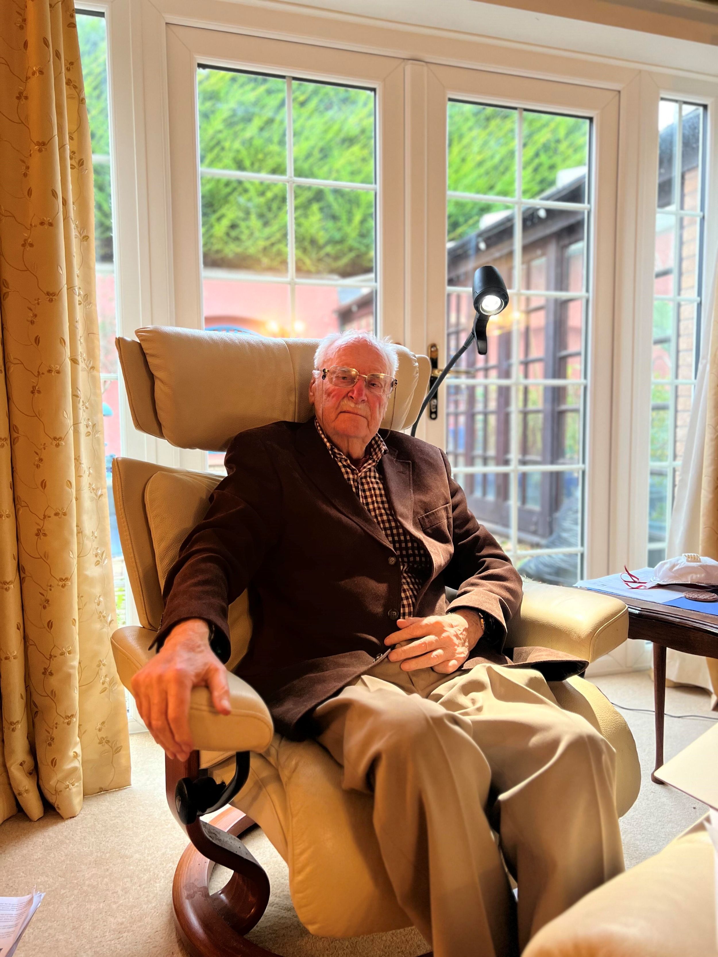 Elderly man wearing glasses, a dark blazer, and checked shirt, sitting in a cream armchair in a living room with large windows behind him.