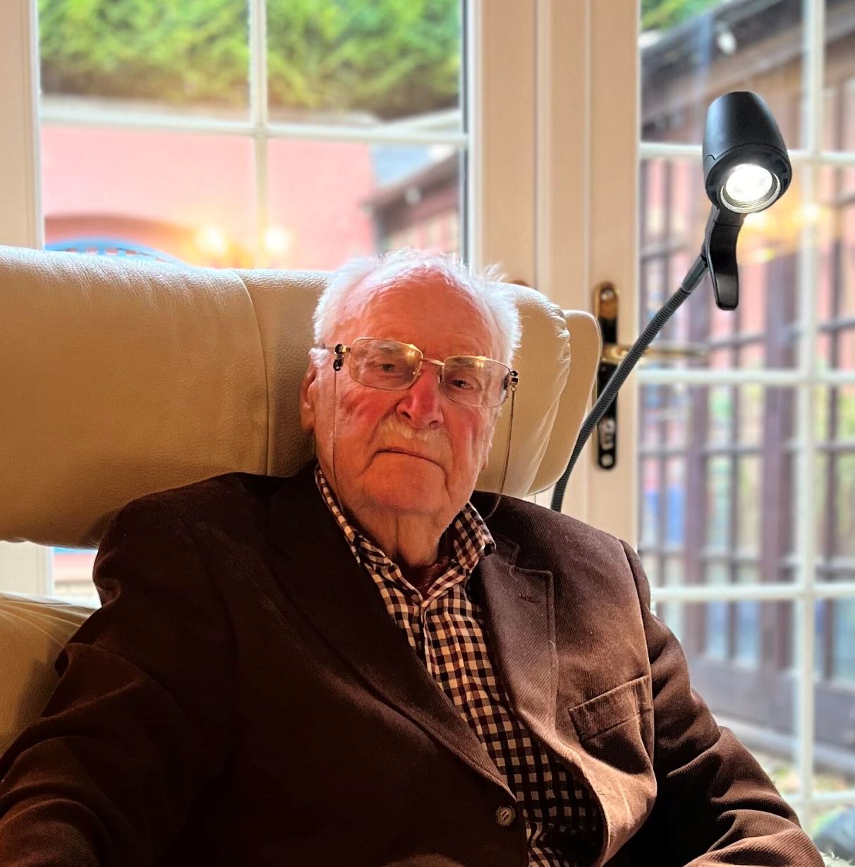 Elderly man wearing glasses, a dark blazer, and checked shirt, sitting in a cream armchair in a living room with large windows behind him.