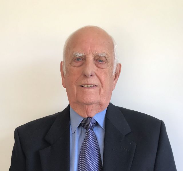 Elderly man with a calm expression, wearing a dark suit jacket, light blue shirt, and patterned tie, standing against a plain background.